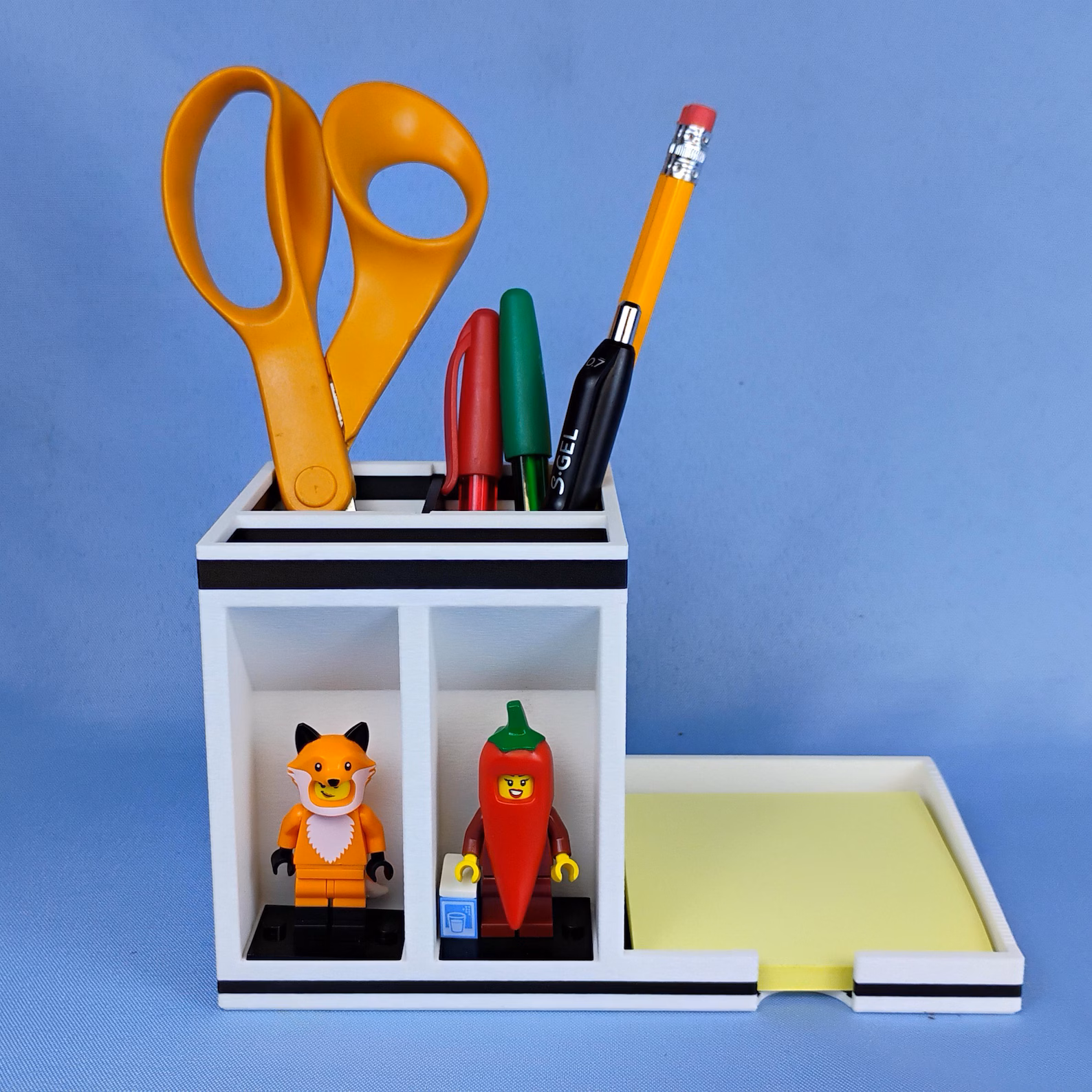 Post-it note holder desk organizer with vertical pen caddy and decorative minifigure display. Provides organized storage for pens, scissors, and sticky notes while adding personality to a workspace.